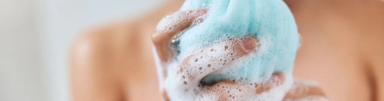 mujer sonriente con un paño exfoliante espumoso en la mano tomando la ducha en casa